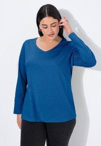 Niebieska koszulka z długim rękawem i dekoltem w serek, wykonana z miękkiego materiału, o luźnym kroju i subtelnej fakturze, zestawiona z ciemnymi dżinsami, przedstawiona na białym tle.