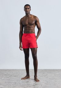 Piros fürdőnadrág rugalmas derékrésszel, húzózsinórral és oldalsó díszítéssel. Simas textúrájú, a comb fölé érő hosszúságú, mezítlábason viselve.