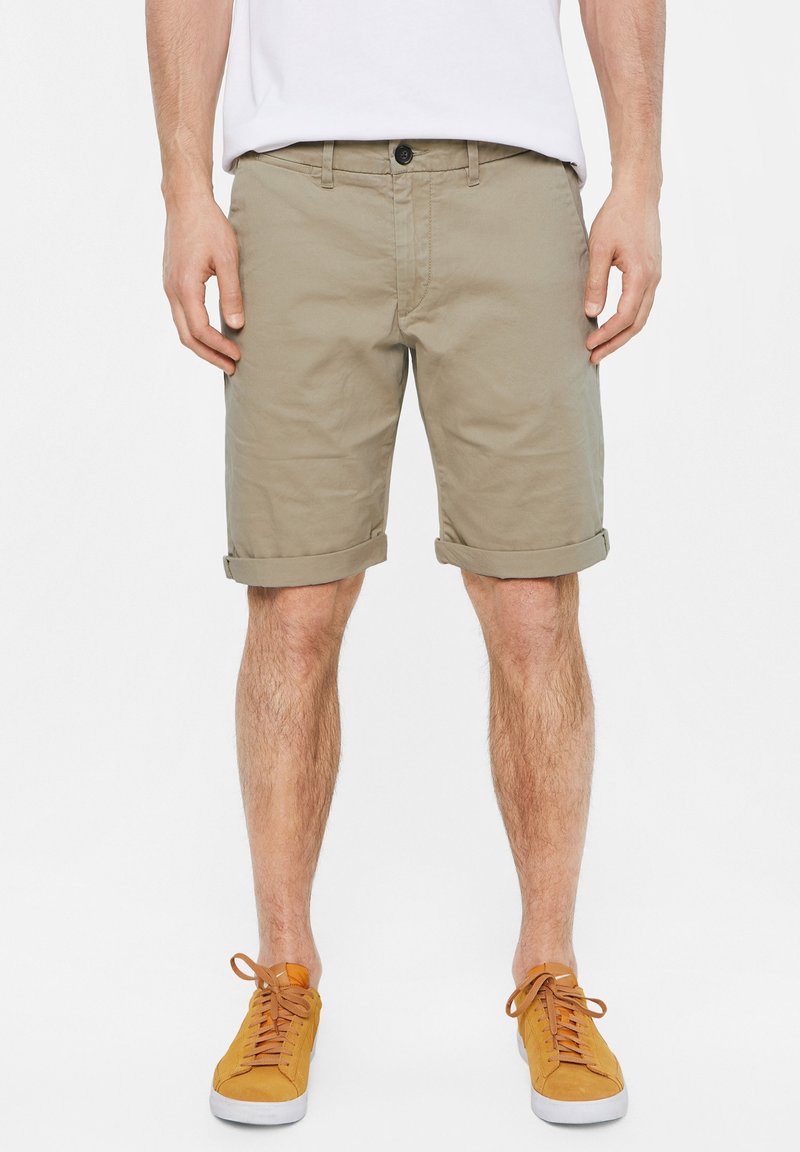 Beige shorts made of cotton, featuring a rolled hem, front pocket, and button closure. Paired with orange sneakers.