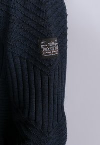 Mörkblå stickad tröja med ett texturerat mönster, med vertikala ribbor och en svart etikett med logotyp och text på axeln.