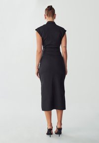 Vestido midi negro sin mangas con una cintura ajustada, aberturas laterales y una textura suave, combinado con zapatos de tacón alto negros.