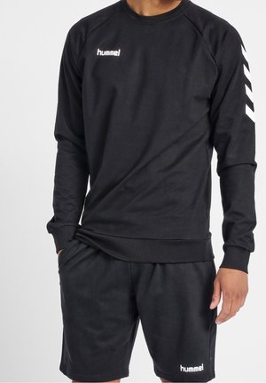 Mand iført sort hummel langærmet sweatshirt og matchende sorte shorts med hummel-logoer på bryst og shorts.