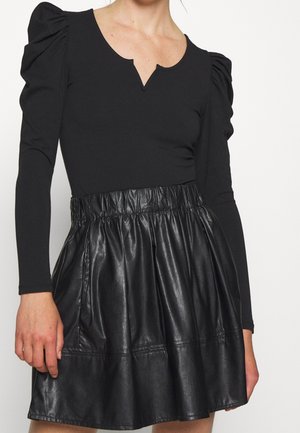 Vrouw die een zwart topje met opgeblazen lange mouwen en een halfronde halslijn draagt, gecombineerd met een hoge taille zwarte rok van nep leer met een geplooide taille.