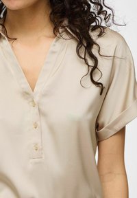 Blusa beige a maniche corte con scollatura a V, dotata di patta e chiusura con bottoni, realizzata in un tessuto leggero e morbido.