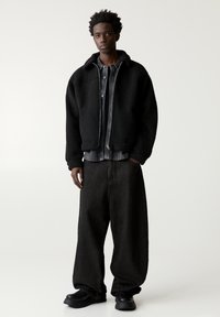 Veste en polaire noire avec fermeture éclair, associée à une chemise en denim foncé et un jean noir ample. Des bottes noires à talons plats complètent le look.