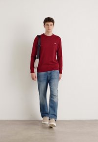 Rød genser, lyseblå jeans, beige joggesko og en grå skulderveske. Genseren har rund hals og figursydd design.