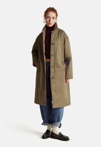 Junge Frau mit lockigem Haar trägt einen langen olivfarbenen Mantel, locker aufgerollte blaue Jeans, weiße Socken, schwarze Loafers und einen dunklen Rollkragenpulli und steht.