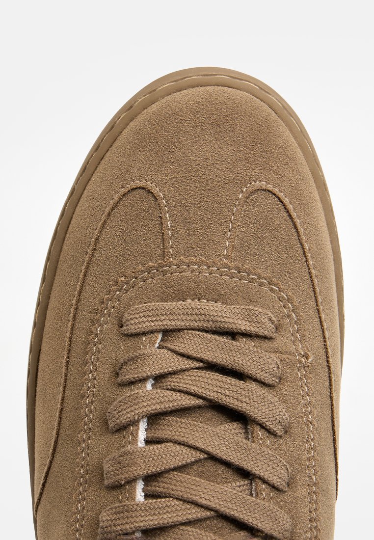 Brązowe zamszowe trampki z zaokrąglonym noskiem, teksturowaną cholewką, przeszyciami oraz płaskimi sznurówkami. Posiadają jasną gumową podeszwę.