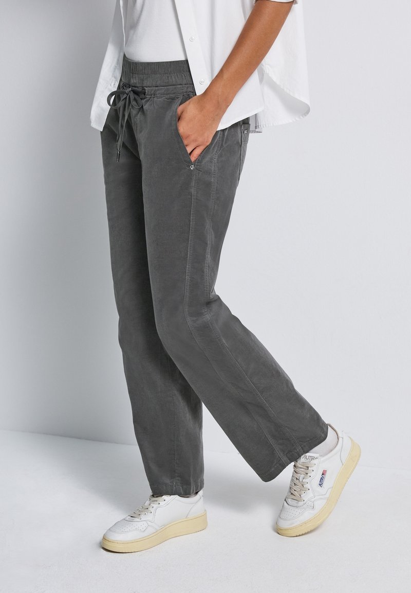 Personne portant un pantalon gris à cordon, des baskets blanches, des chaussettes blanches et une chemise blanche partiellement visible, debout devant un fond gris clair.