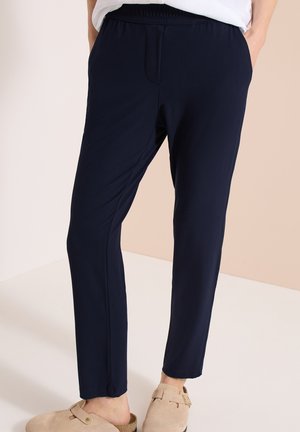Person iført marineblå slim-fit bukser med elastisk talje og beige slip-on sko, stående mod en lys baggrund.