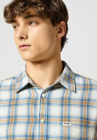 Geruite overhemd in beige, blauwe en witte tinten. Voorzien van een button-down kraag, twee zichtbare knopen en een klein "Wrangler" logo op de borst.