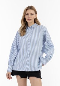 Blå og hvid stribet langærmet skjorte med knaplukning, afslappet pasform og manchetter, båret over sorte slidte shorts.