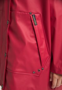 Giacca impermeabile rossa brillante con una superficie liscia, chiusure a scatto, una grande tasca inclinata e un'etichetta con logo sulla tasca.