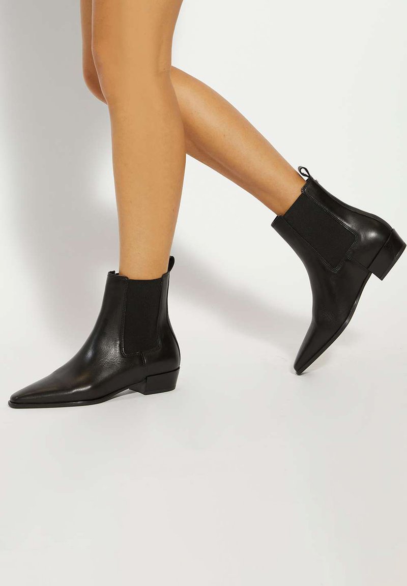 Jambes portant des bottines en cuir noir élégantes avec des bouts pointus et des talons blocs bas sur un fond clair uni.