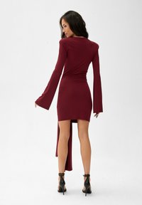 Burgundské dlouhé šaty s upnutým tělem a vysokým rozparkem. Hladká látka a široké rukávy vytvářejí elegantní siluetu. Černé vysoké podpatky.