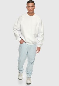Sweatshirt blanc surdimensionné avec des épaules tombantes, associé à un jean en denim bleu clair. Les chaussures ont une base blanche avec des accents jaunes.
