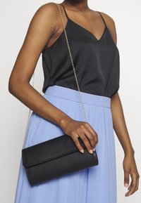 Černá kabelka z umělé kůže s přehnutým vrškem a zlatým řetízkovým popruhem, kterou drží osoba oblečená v černém topu a pastelově modré sukni.