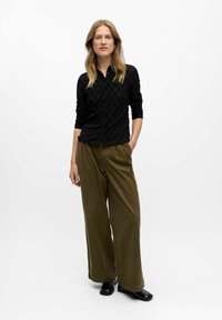 Haut en tricot noir à boutons avec un motif texturé, associé à un pantalon large vert olive décontracté et des chaussures habillées noires.