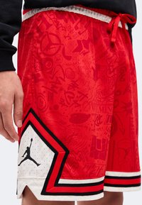 Κόκκινα shorts μπάσκετ με διακριτό γραφικό σχέδιο, μαύρες και άσπρες λεπτομέρειες, κορδόνι στη μέση και μαύρο λογότυπο Jumpman κοντά στο τελείωμα.