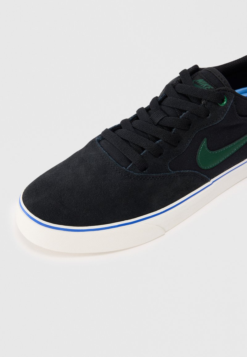 Czarne zamszowe sneakersy z zielonym logo Nike, niebieskimi akcentami na podeszwie i czarnymi sznurówkami. Posiadają białą gumową podeszwę.