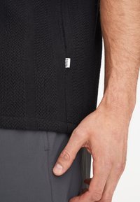 Pullover nero in maglia testurizzata con un motivo a coste. Presenta un piccolo etichetta bianca del marchio all'orlo. Pantaloni grigi visibili in basso.