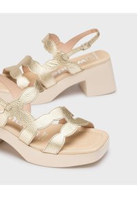 Sandalias trenzadas de color dorado con un diseño texturizado, que cuentan con un tacón grueso beige y una correa en el tobillo ajustable. Plantilla suave con el logo.