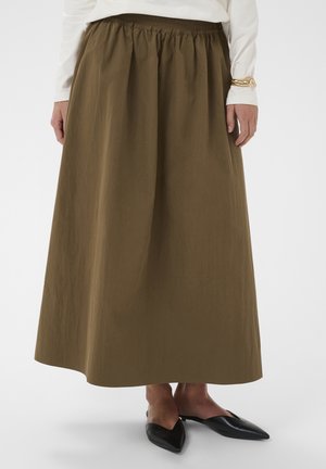 Naine, kes kannab pruuni pikka seelikut, valget pika varrukaga pluusi, kuldset käevõru ning musti teravnurkadega madalaid kingi, seisab valgel taustal.