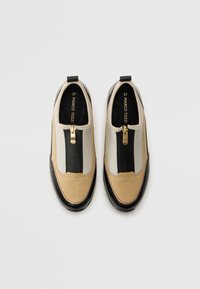 Un paio di scarpe slip-on beige, nere e bianche con chiusure a zip dorate e marchio "Marco Tozzi" all'interno e sui lati.