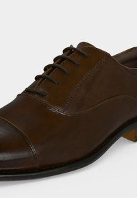 Chaussure habillée à lacets en cuir marron, avec bout droit et coutures apparentes, montrée en gros plan de côté sur un fond neutre.
