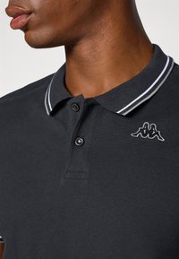 Svart polo skjorte med teksturert stoff, med en hvit stripet krage og knapplist med tre knapper. Kappa-logo brodert på brystet.