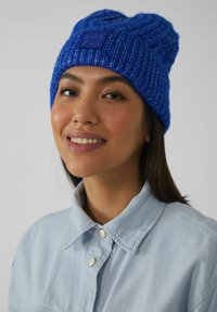 Blauwe gebreide muts met geribbelde textuur en een tonale logo-patch. Draagt bij een lichtblauw overhemd met een kraag, dat snap-knopen heeft.