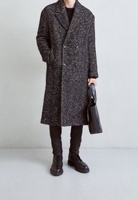 Osoba v dlouhém kabátě s černo-bílým vzorem, černých kalhotách, černých botách, držící černou koženou kabelku před jednobarevnou světlou zdí.