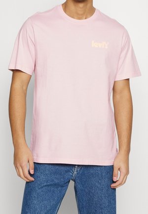 Mężczyzna w jasnoróżowej koszulce Levis i niebieskich dżinsach, stojący z rękami swobodnie opuszczonymi wzdłuż ciała na jednolitym tle.