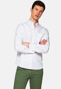 Biała koszula z guzikami, z małym niebieskim kwiatowym wzorem, długie rękawy i mankiety. Połączona z zielonymi spodniami dla kontrastowego wyglądu.