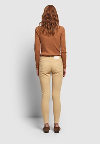 Mujer con cabello ondulado rojo, vistiendo un suéter acanalado color óxido, pantalones de piel ajustados beige y mocasines de estampado de leopardo, de pie contra un fondo liso.