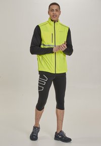 Helle gelbe ärmellose Jacke mit hohem Kragen, reflektierendem Streifen und langen schwarzen Ärmeln. Schwarze enganliegende Leggings mit einem weißen grafischen Muster.