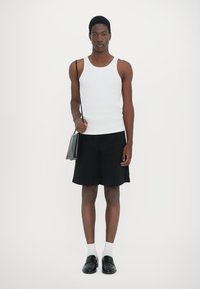 Débardeur blanc à côtes, shorts noirs, mocassins noirs, chaussettes blanches arrivant aux chevilles, et un petit sac noir. Design simple et couleurs neutres.