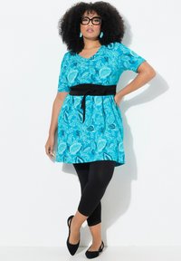 Turkusowa sukienka w kwiaty z krótkimi rękawami i czarnym paskiem, w połączeniu z czarnymi legginsami i czarnymi butami, z dekoltem w serek i plisowanym wzorem.