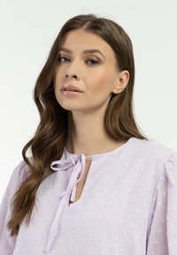 Světle fialová blůza s texturovanými puntíky, kulatým výstřihem a detailem v podobě mašle. Rukávy jsou mírně nabírané, což dodává designu objem.