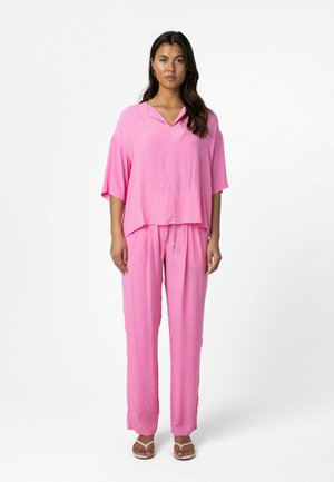 Vrouw staande, gekleed in een losse roze V-hals top met korte mouwen en bijpassende broek met trekkoord, en witte sandalen met hakken, op een witte achtergrond.