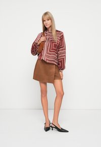 Brauner Wildlederrock kombiniert mit einer bordeauxfarbenen gemusterten Bluse mit geometrischen Streifen; Modell hält eine kleine braune Tasche; schwarze spitze Absätze.