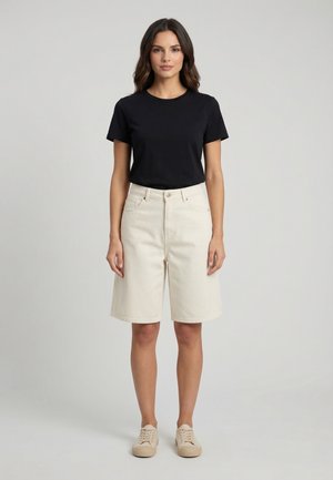 Femme debout de face, portant un t-shirt noir à manches courtes, un short écru jusqu'aux genoux et des baskets beige, sur un fond uni.