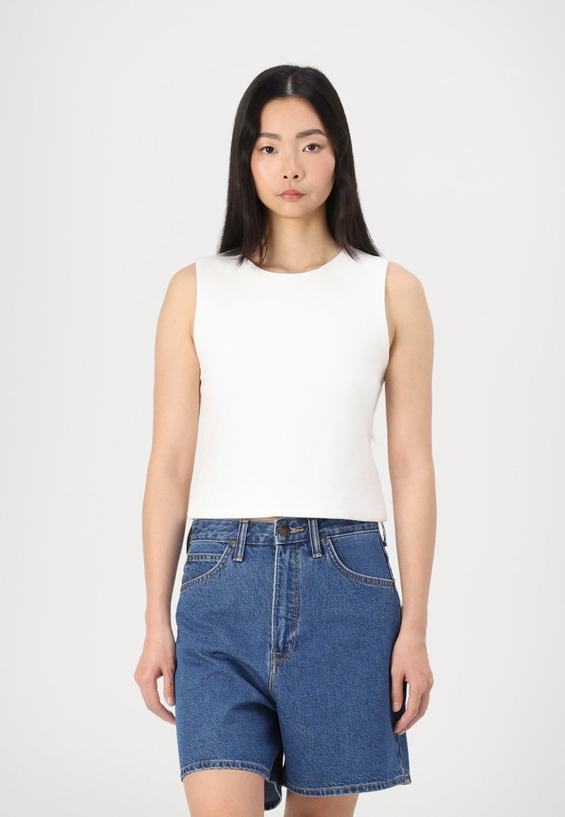 Top blanc sans manches avec un col rond associé à un short en denim bleu taille haute, comportant deux poches avant et des coutures visibles.