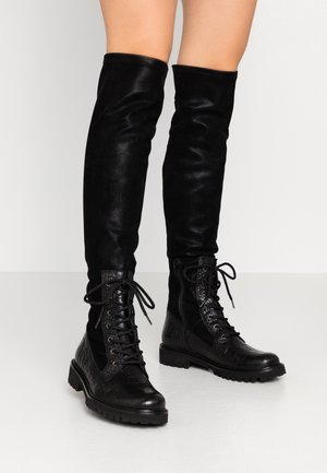 Zwarte kniehoge laarzen met veters aan de voorkant, een getextureerde leren neus en een stevige zool, gedragen op blote benen tegen een effen witte achtergrond.
