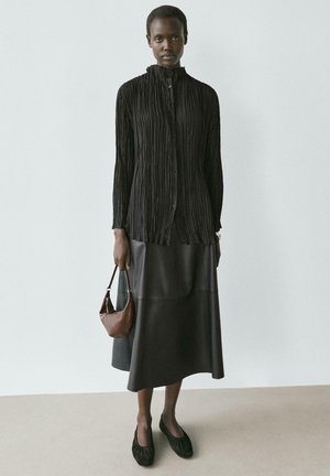 Chemise plissée noire à manches longues, boutonnée, associée à une jupe droite en cuir noir. Un sac à main marron et des chaussures noires sans lacets complètent le look.