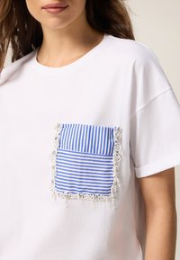 T-shirt bianco con tasca a righe blu caratterizzata da dettagli texturizzati e piccoli accenti decorativi lungo i bordi. Maniche corte.