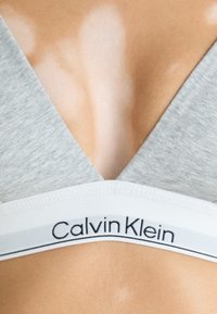 Sivi pamučni bralet s dizajnom u obliku slova V. Ima bijelu elastičnu traku s logotipom "Calvin Klein" i suptilnom teksturom.