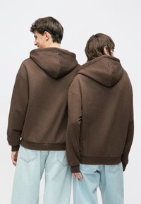 Braune Kapuzenpullover mit gerippten Bündchen an Ärmeln und Bund, gezeigt von hinten an zwei Personen, kombiniert mit hellblauen, lockeren Jeans.