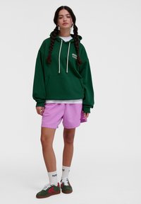 Green oversized hoodie with drawstrings and front pocket, paired with pink shorts. Footwear includes green shoes with red accents and white socks.