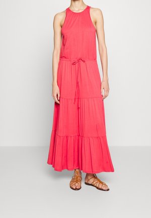 Kobieta w czerwonej maxi sukience bez rękawów z wiązanym w pasie i falbanami na spódnicy, w połączeniu z brązowymi sandałami na płaskiej podeszwie z paskami.